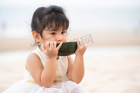 沙滩上的孩子摄影照片_可爱的孩子吃西瓜在热带沙滩在夏天。可爱的孩子在沙滩上吃西瓜。