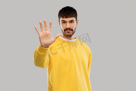 黄色手指手势摄影照片_警告和人的概念—严肃的年轻人在黄色运动衫显示停止手势在灰色背景。严肃的年轻人显示停止手势