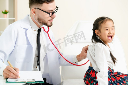 孩子住院摄影照片_年轻的男医生在医院办公室检查小孩。孩子很高兴，不怕医生。儿童医疗保健概念。