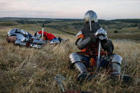 中世纪的骑士，穿着盔甲，戴着头盔，在伟大的战斗后坐在地上。身穿盔甲的古代战士在田野中摆姿势