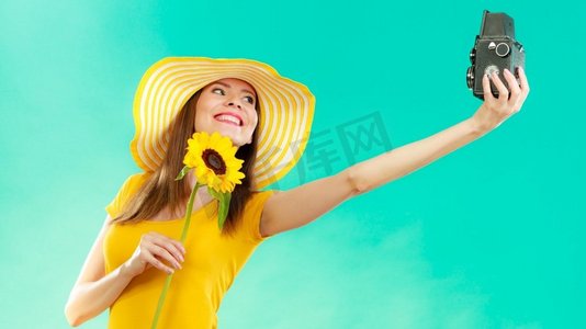 黄色照片摄影照片_一位身穿黄色连衣裙、戴着向日葵帽子的夏日女子在鲜艳的蓝色背景下用老式相机自拍
