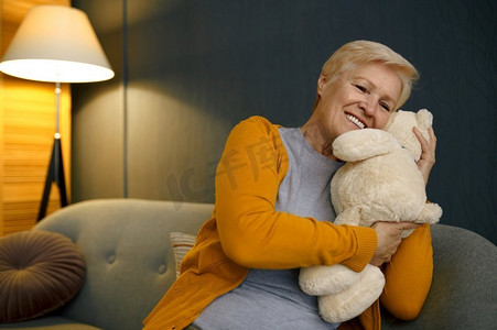 老妇人抱着泰迪熊坐在沙发上思念孙子和长期分离。老妇人拥抱泰迪熊在沙发上