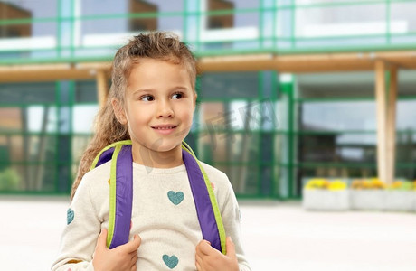 童年和教育理念-快乐的小女孩背着背包盖在学校院子里。背着书包的快乐小女孩