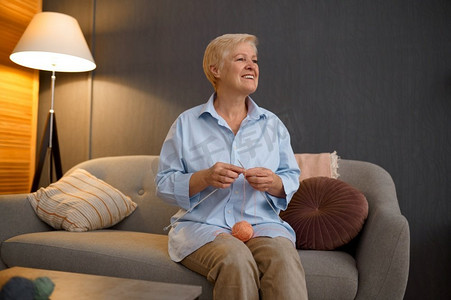 祖母编织与纱球坐在家沙发上。退休概念中的手工艺爱好。微笑的祖母编织坐在家里沙发