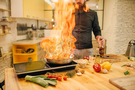 火焰摄影照片_男厨师用火烧肉，火焰进入煎锅，厨房为背景。一名男子在桌上电炉上准备牛肉