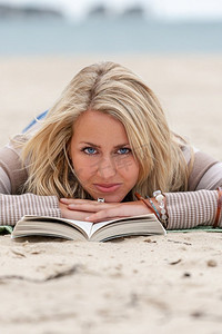 有蓝色眼睛和金发的美丽的年轻女子女孩躺下阅读平装书在海滩