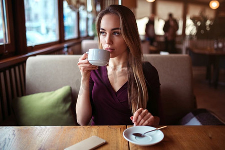 喝饮料的女孩摄影照片_一名年轻女子在咖啡馆喝饮料。美丽的女孩端着一杯咖啡，看着餐厅的窗户。