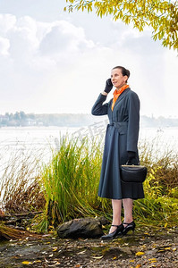 优雅的资深女商人，拿着手机，背着包，戴着橙色的围巾，在秋天的河边散步，在树下