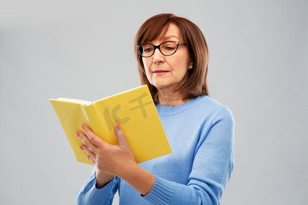 老人阅读摄影照片_视野、智慧和老人观念--灰色背景下戴眼镜看书的资深女性。戴眼镜的资深女性阅读书籍