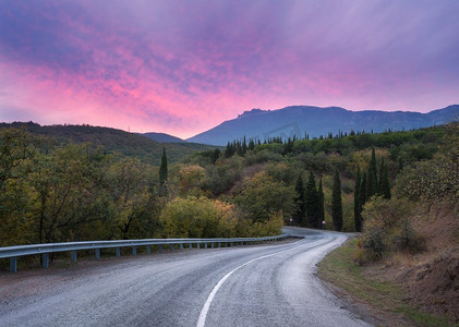 山蜿蜒的道路通过森林与五颜六色的天空和粉红色的云在日落在夏天