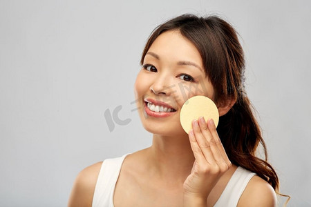 美容，人和护肤概念—年轻的亚洲妇女清洁面部与去角质海绵。亚洲妇女清洁脸与去角质海绵