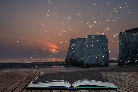 萤火虫之墓摄影照片_在岩石栈的美丽的五颜六色的日落在海滩风景与萤火虫发光在幻想样式的图像出来的页打开的故事书