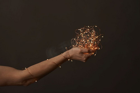 圣诞花环摄影照片_一个女人的手裹着圣诞花环，在黑暗的背景下手里拿着一把火。’明亮的圣诞灯在妇女的手中’