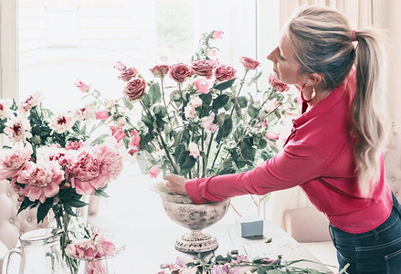 穿着红色衬衫的花店妇女，在窗边的桌子上用金塔花瓶里的玫瑰和其他鲜花制作美丽的大型节日活动的古典花束，生活方式