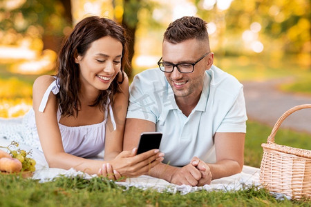 手机夏季夏季摄影照片_休闲和人的概念—快乐的夫妇与智能手机有野餐在夏季公园。愉快的夫妇与智能手机在野餐在公园