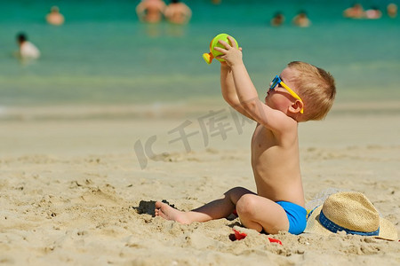 沙滩上的孩子摄影照片_两岁的孩子在沙滩上玩耍。两岁的蹒跚学步的男孩在海滩上玩沙滩玩具 
