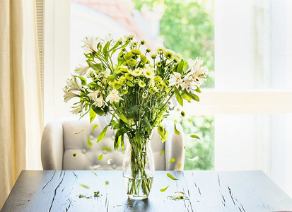 美丽的绿色花束与落花瓣在玻璃花瓶表在阳光明媚的客厅在窗口。家居内饰和装饰理念