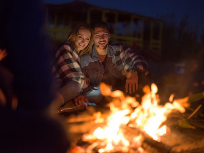 年轻夫妇与朋友坐在篝火周围在海滩晚上喝啤酒。夫妇享受与朋友在晚上在海滩上