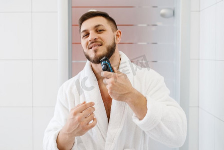 身穿浴袍的男子在浴室里用电动剃须刀刮胡子，这是例行的早晨卫生。一名男子用电动剃刀刮胡子