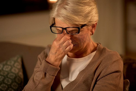 老年，视力和人的概念—累了的高级妇女在眼镜擦她的眼睛在家里晚上。晚上在家里戴眼镜的疲惫的高级妇女