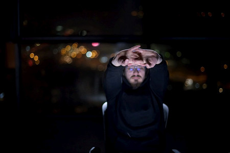 大城市摄影照片_年轻的天才黑客在黑暗的办公室里工作时使用笔记本电脑，晚上背景是大城市的灯光