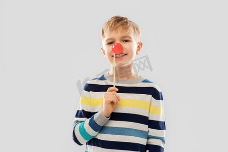 红鼻子日，照相亭和童年概念-灰色背景下带着小丑鼻子的微笑小男孩。戴着红色小丑鼻子的微笑男孩派对道具