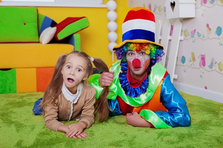 微笑的孩子和小丑躺在婴儿室的地毯上。彩色沙发，嵌套盒和栅栏在背景。 