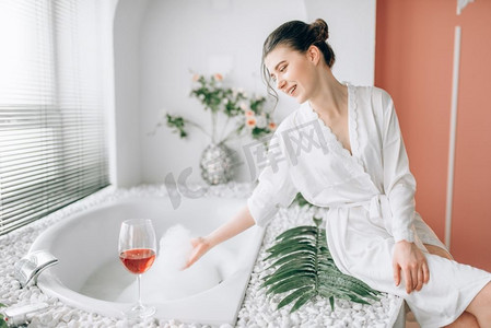 在白色浴衣的年轻妇女坐在与泡沫的浴缸边缘。浴室内部与窗口和玻璃与红葡萄酒在背景