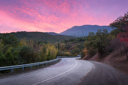 山蜿蜒的道路通过森林与戏剧性的五颜六色的天空和红色的云彩在日落在夏天