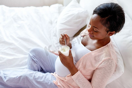 怀孕、人和食物概念-愉快的怀孕的吃酸奶的非洲裔美国妇女在床上在家孕妇在床上吃酸奶