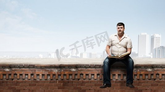 胖子坐在建筑物顶部与书。胖子