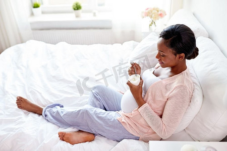 怀孕、人和食物概念-愉快的怀孕的吃酸奶的非洲裔美国妇女在床上在家孕妇在床上吃酸奶