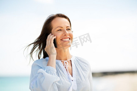 技术，人和休闲概念—愉快的微笑的妇女呼叫智能手机在夏季海滩。快乐的女人呼叫智能手机上夏季海滩