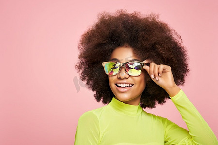 在彩色全息太阳镜的非洲裔美国人女孩的美丽肖像。美丽的黑人妇女在粉红色背景。化妆品、化妆品和时尚