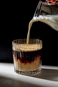 一杯牛奶杯摄影照片_咖啡因和饮料的概念—用壶把奶油或牛奶倒进桌上的一杯咖啡里。手拿着壶把奶油倒进一杯咖啡里
