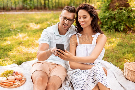 手机夏季夏季摄影照片_休闲与人的概念快乐夫妇带着智能手机在夏季公园野餐。带着智能手机在公园野餐的幸福情侣