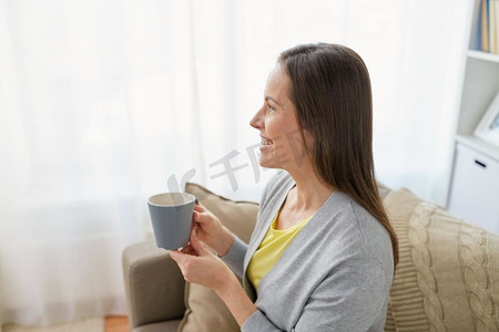 女人酒摄影照片_人、酒和休闲的概念--快乐的中年妇女端着杯子在家喝茶或喝咖啡。快乐的女人在家喝茶或喝咖啡