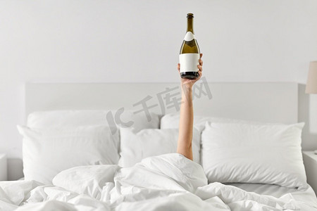 女人酒摄影照片_酒精，舒适和早晨概念—年轻妇女的手躺在床上与香槟瓶。女人的手躺在床上与香槟