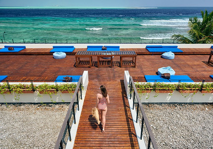 连体比基尼摄影照片_穿着红色条纹连体式泳衣的女子走在热带海滩露台甲板上。在马尔代夫度暑假。