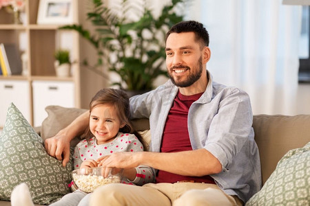 亲和摄影照片_家庭，父亲和人的概念—快乐的父亲和女儿与爆米花在家里看电视。快乐的父亲和女儿在家里看电视