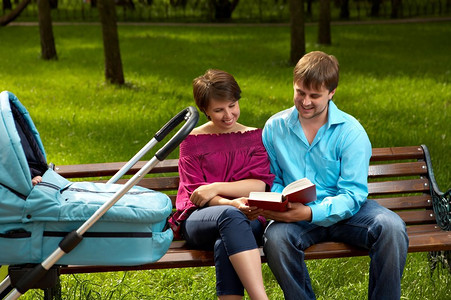 已婚夫妇在散步与婴儿坐在公园的长凳上，并在草坪的背景上阅读这本书。家庭步行