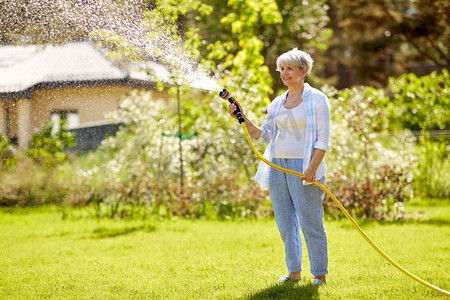 园艺和人的概念—快乐的高级妇女浇水草坪由花园软管在夏天。高级妇女浇水草坪由软管在花园