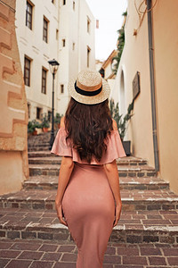 享受旅行摄影照片_美丽的深色头发的年轻女子穿着粉色连衣裙，戴着草帽走在欧洲老城的街道上。时尚和风格。暑期旅行