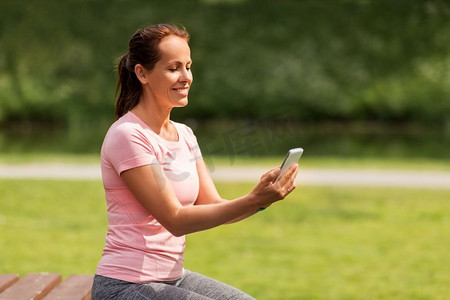 健身，运动和健康的生活方式概念—快乐的微笑的妇女与智能手机在夏天公园。女人与智能手机在夏季公园