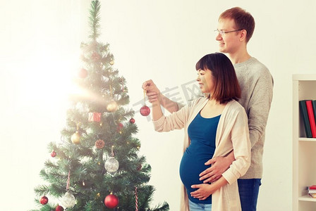 怀孕，寒假和人们概念幸福的夫妇在家里装饰圣诞树。圣诞树旁快乐的怀孕夫妇和丈夫