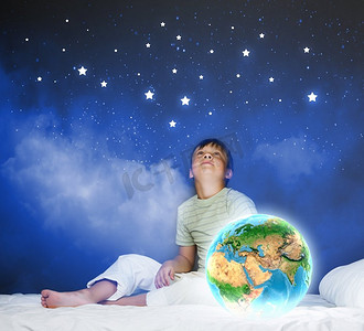 夜里做梦。可爱的男孩坐在床上看着地球。这张图片的要素由美国宇航局提供