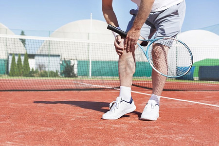 夏季红色网球场上的成熟男子拿着网球拍而遭受膝盖疼痛