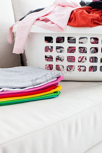叠衣服摄影照片_在篮子里洗衣服，在沙发上放一叠干净的亚麻布。五颜六色的干净衣服