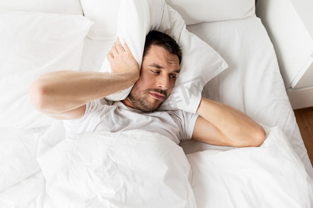 古人枕头摄影照片_人，睡觉时间和休息的概念-男人躺在床上枕头在家受噪音困扰。躺在床上枕头受噪音折磨的男人