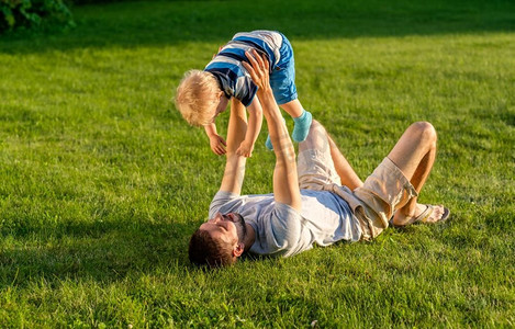 家庭场景摄影照片_快乐的男人和孩子在草地上享受户外乐趣。公园里父子俩在绿茵茵的草地上休息的家庭生活场景。